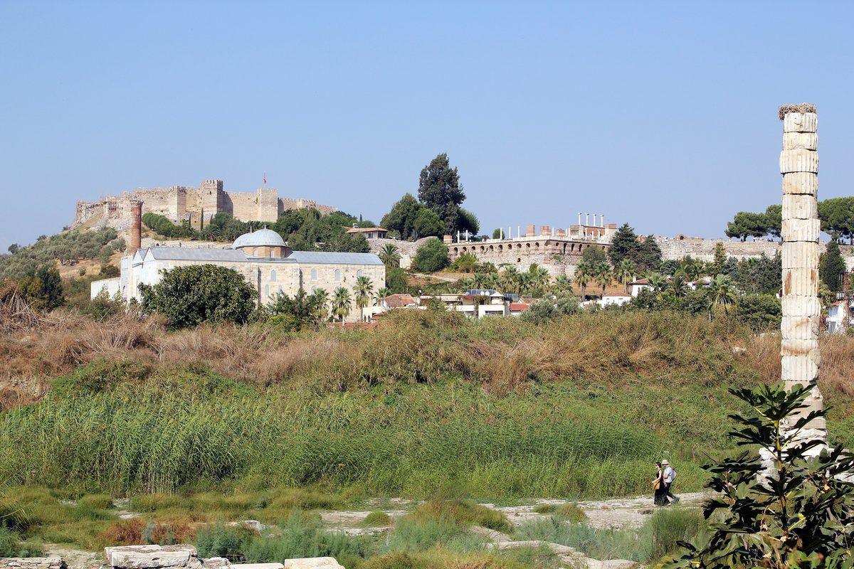Vista del solar del Templo de Artemisa entre juncos y hierba alta, con dos visitantes en el sendero y la columna a la derecha; al fondo la Mezquita de Isa Bey, la Basílica de San Juan y la fortaleza de Ayasuluk