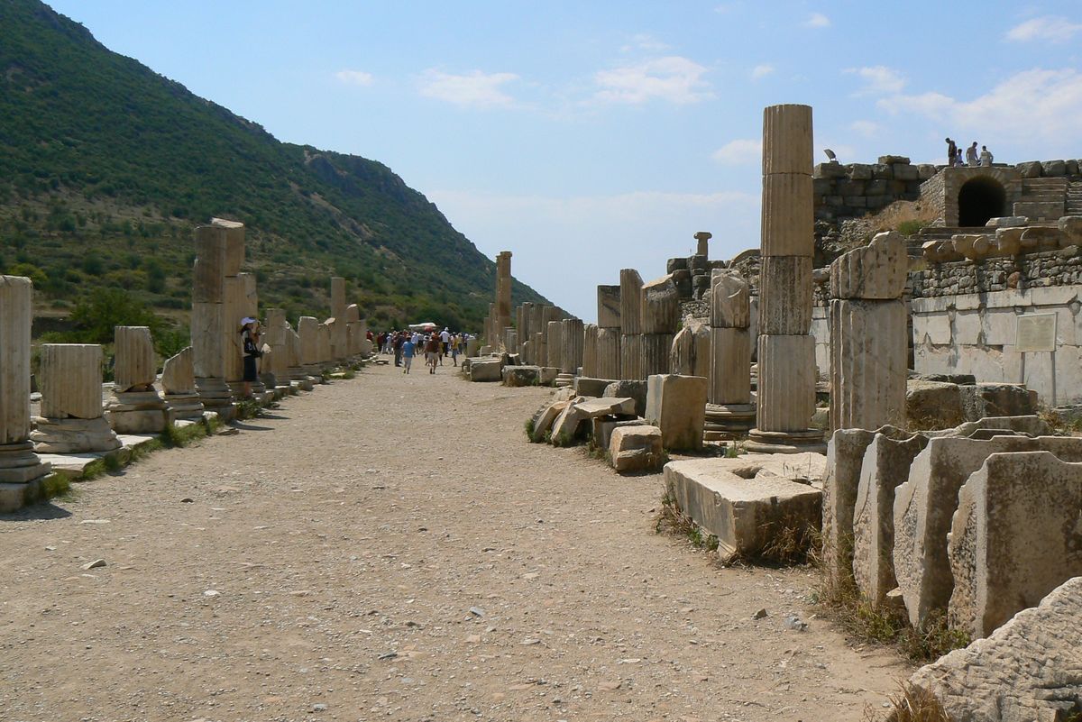 Fustes de columna a ambos lados de un camino de tierra en la parte alta de Éfeso, donde estuvo la Basílica Estoa del ágora de Estado, con la ladera verde del monte a la izquierda