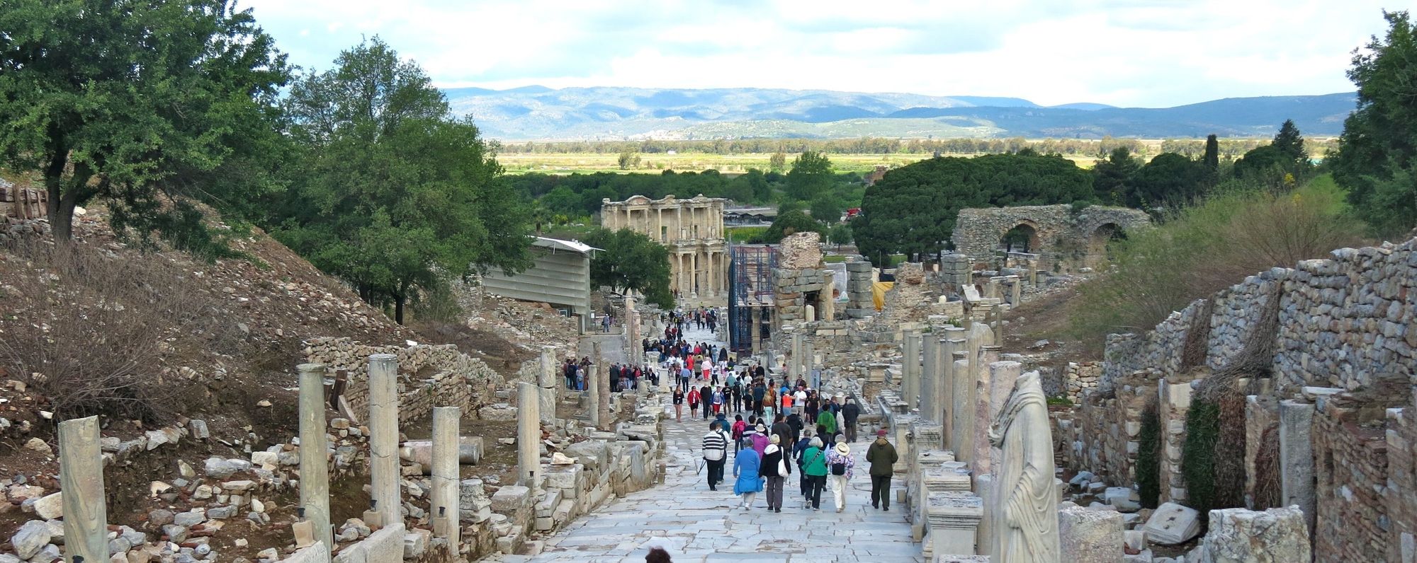 La calle de los Curetes de Éfeso vista cuesta abajo: una vía de losas de mármol entre columnas y estatuas sin cabeza, llena de visitantes de espaldas, con la fachada de la Biblioteca de Celso al fondo y la llanura y los montes detrás