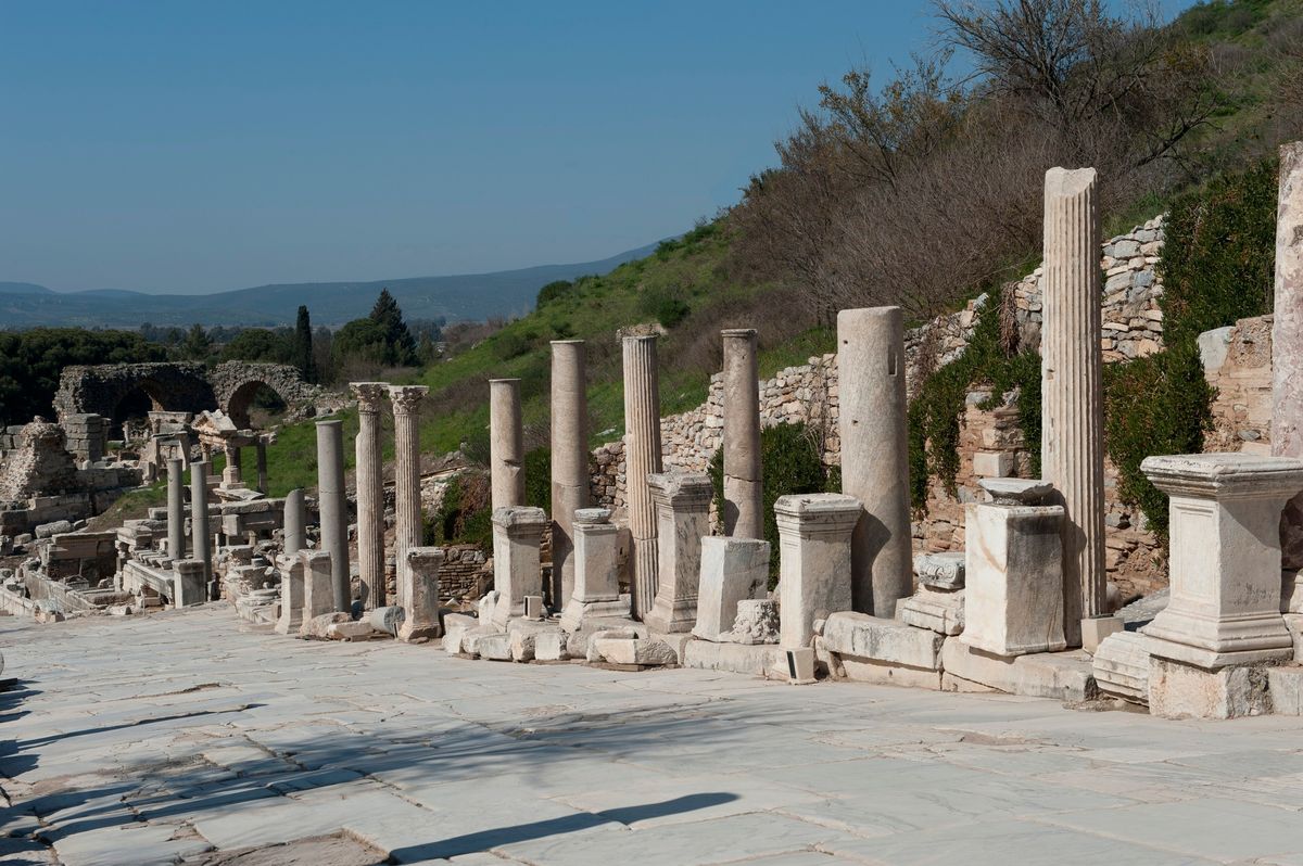 La calle de los Curetes de Éfeso cuesta abajo, pavimentada de mármol, con una hilera de columnas y pedestales de estatuas a la derecha y la ladera verde al fondo