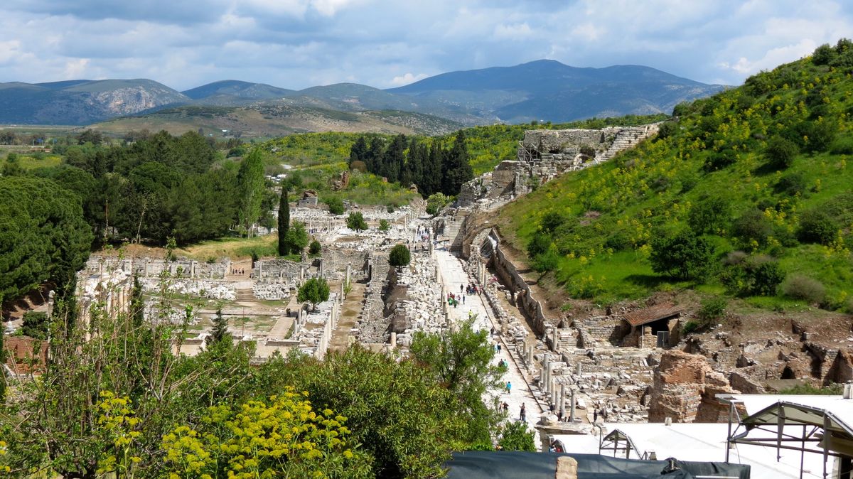 Vista desde la ladera de Éfeso: a la izquierda las ruinas del ágora comercial, en el centro una calle de losas con visitantes que avanza hacia el Gran Teatro, excavado en la colina de la derecha