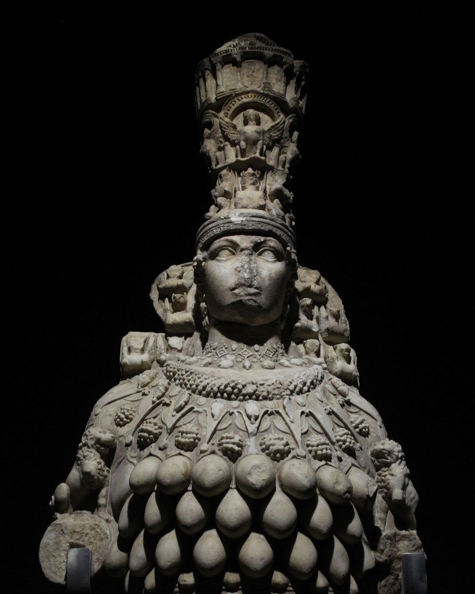 Estatua de mármol de la Artemisa de Éfeso sobre fondo negro: corona alta con un templo en miniatura, collares, leones en los brazos y el pecho cubierto de formas ovaladas