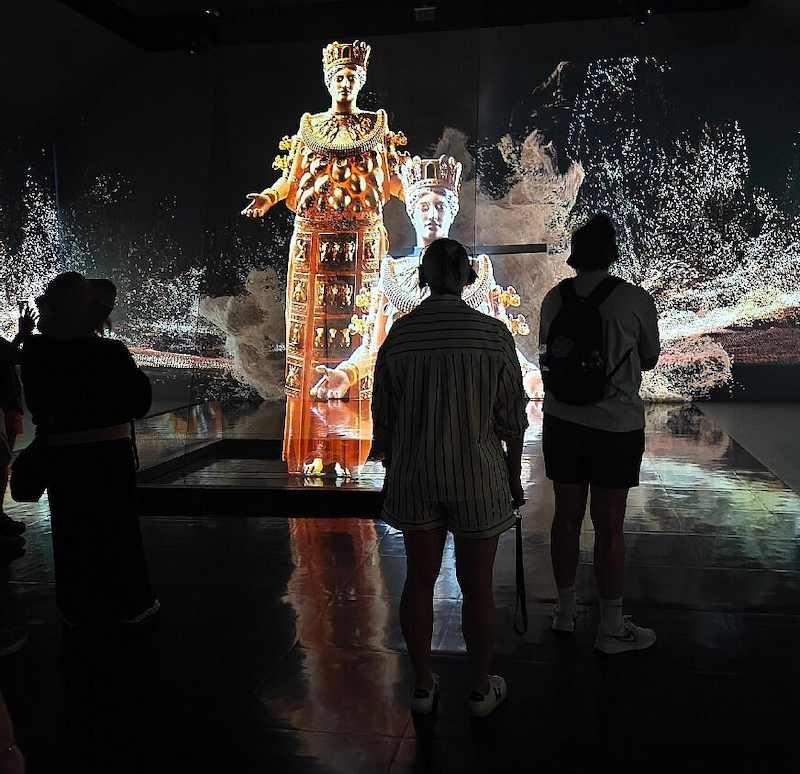 Tres visitantes de espaldas en una sala oscura del Museo de la Experiencia de Éfeso, ante la figura iluminada de la Artemisa de Éfeso rodeada de proyecciones