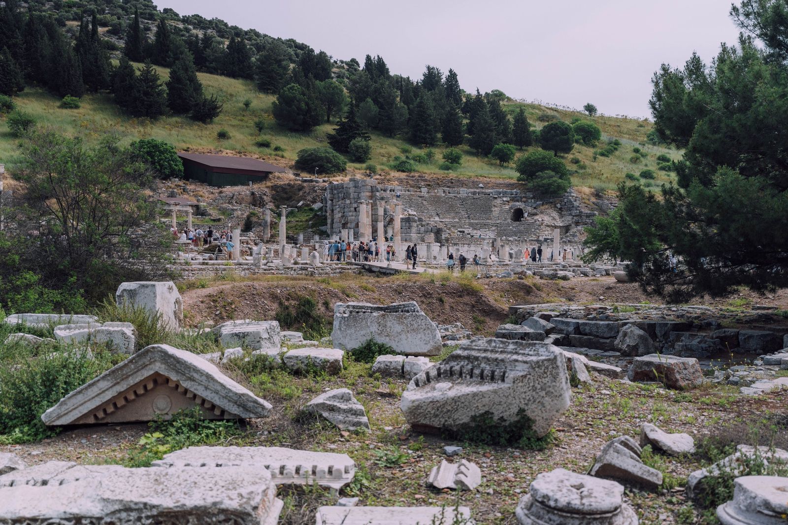 Bloques de cornisa tallados en primer plano y, al fondo, las columnas de la Basílica Estoa con grupos de visitantes delante de las gradas semicirculares del odeón de Éfeso, al pie de una ladera con pinos y cipreses