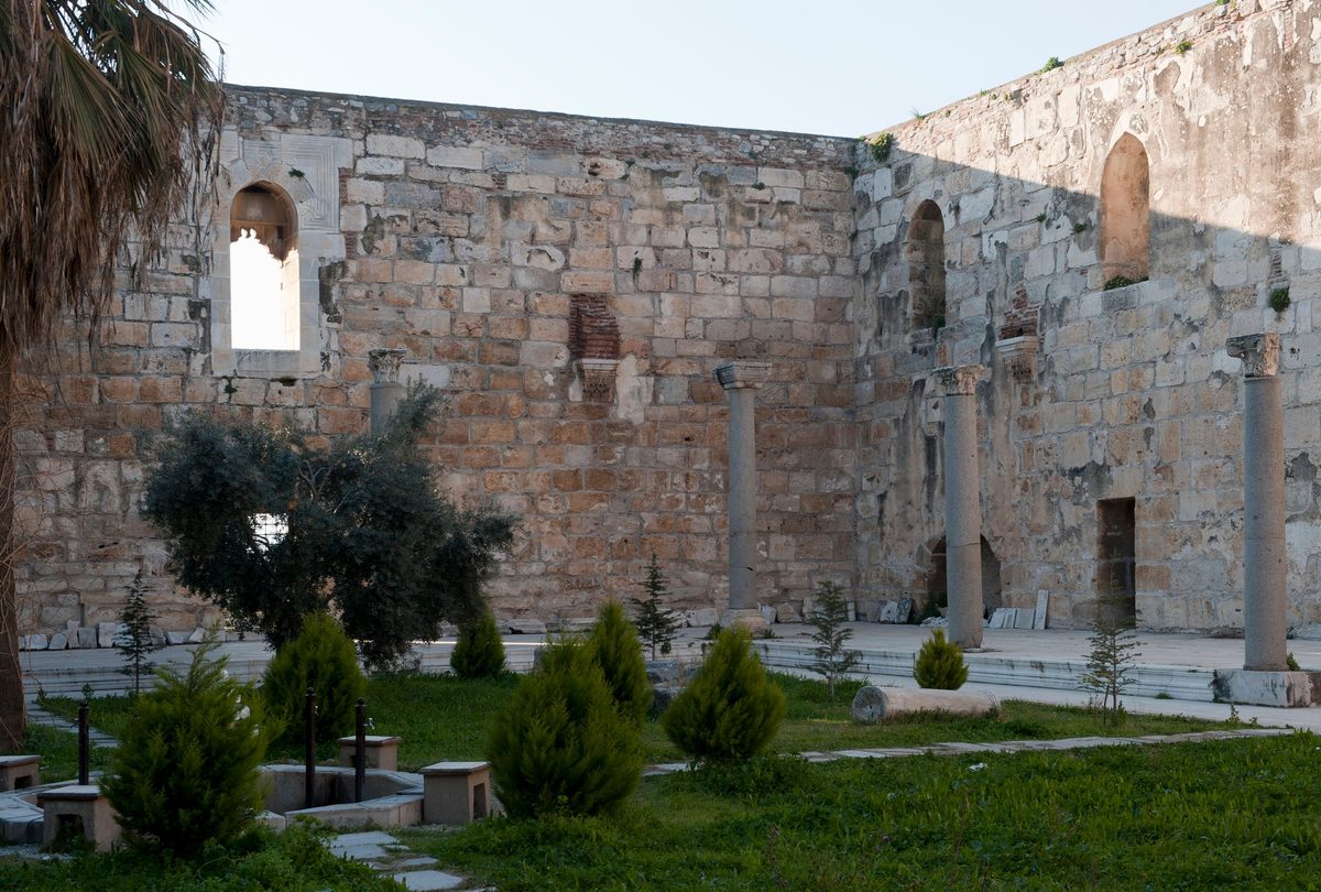 Patio de la Mezquita de Isa Bey en Selçuk: altos muros de sillares de piedra reaprovechada, ventanas con arcos apuntados y columnas antiguas de granito, con cipreses y un jardín delante