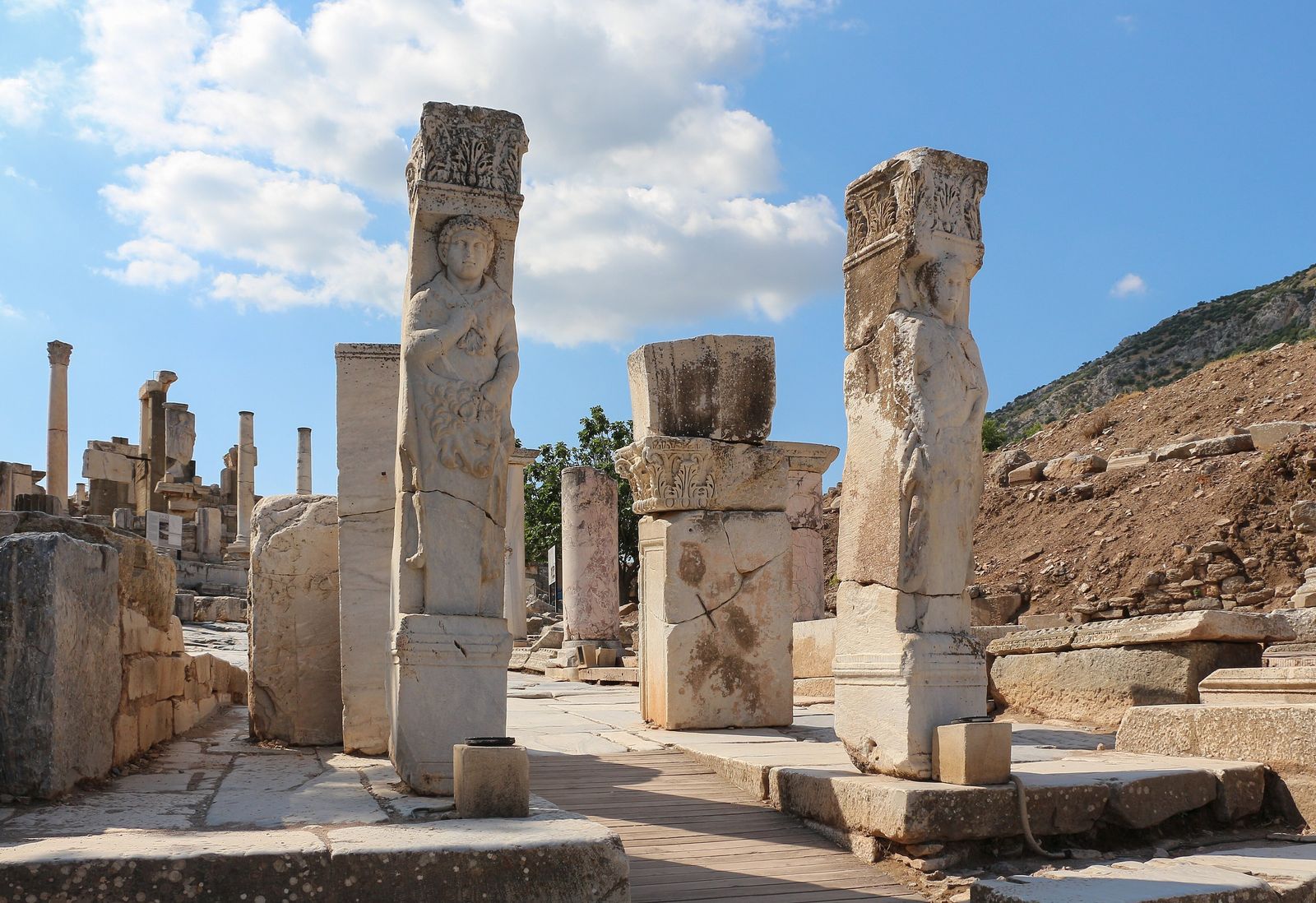 Los dos pilares de mármol de la puerta de Hércules en Éfeso, uno con el relieve de Hércules cubierto con la piel de león, sobre una pasarela de madera que baja por la calle de los Curetes entre columnas y bloques bajo un cielo con nubes