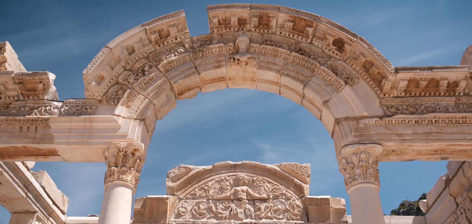 El arco del Templo de Adriano en Éfeso visto desde abajo, con un busto femenino en la clave y, detrás, una figura con los brazos abiertos entre hojas de acanto sobre la puerta