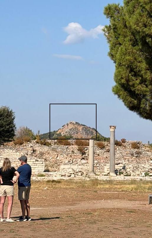 Colina al oeste de Éfeso con la torre de las antiguas murallas en la cima, señalada con un recuadro negro, vista desde las ruinas con dos columnas y una pareja de visitantes de espaldas