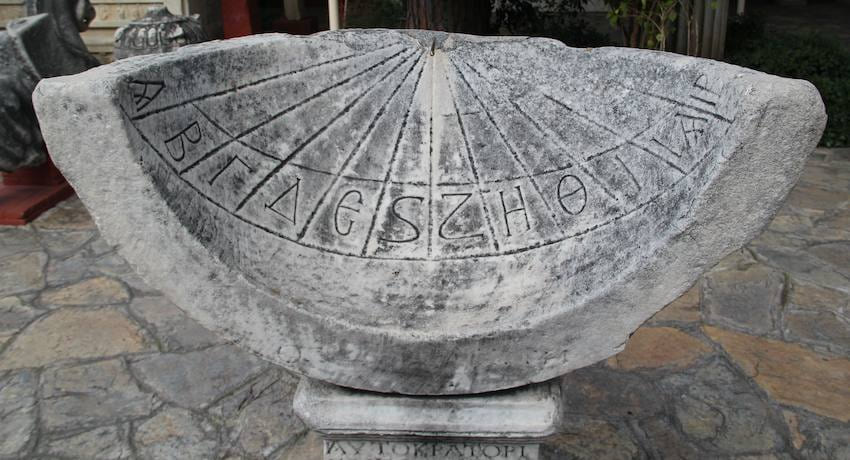 Reloj de sol antiguo de mármol con líneas horarias y letras griegas talladas, sobre un pedestal de piedra